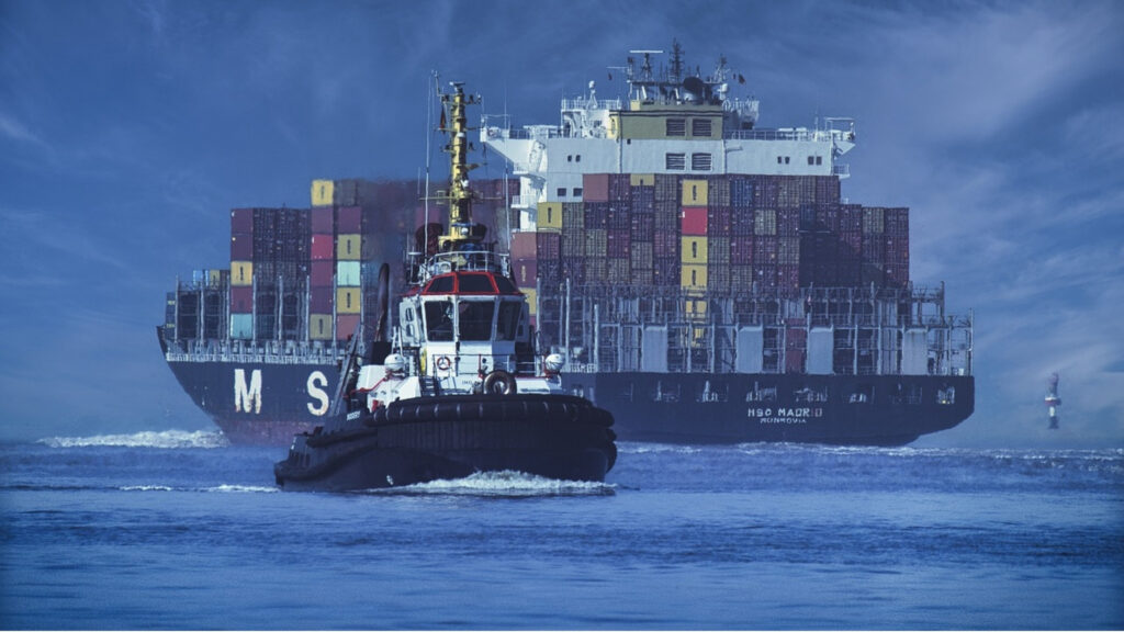 Cargo ship carrying large stacks of containers being pulled by a tug boat