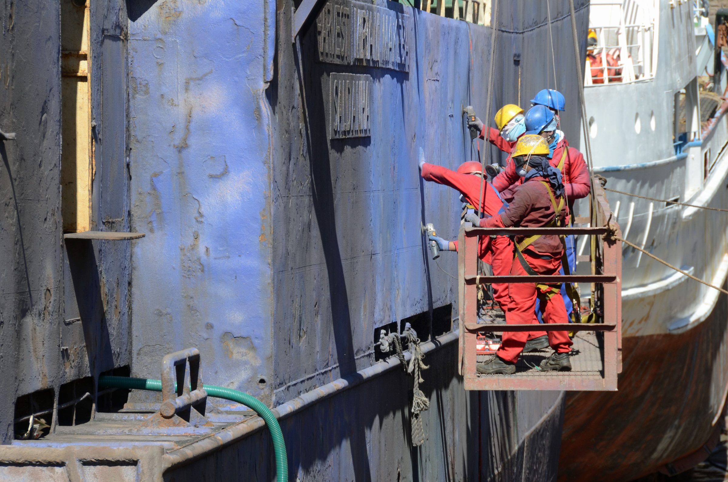 A crew in coveralls and harnesses working a ship hull from a suspended cradle
