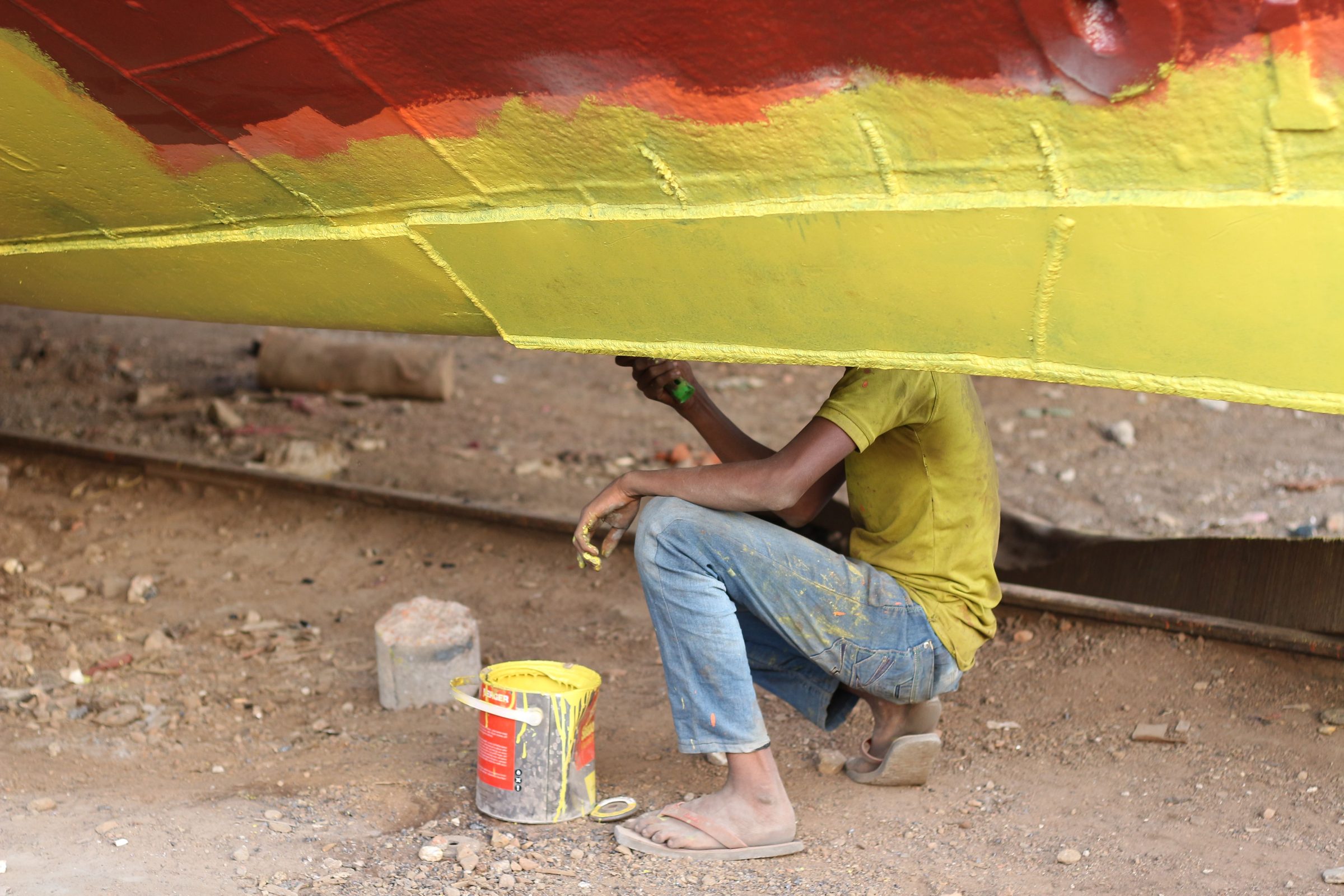 A worker painting the underside of a hull with a paint tin beside him