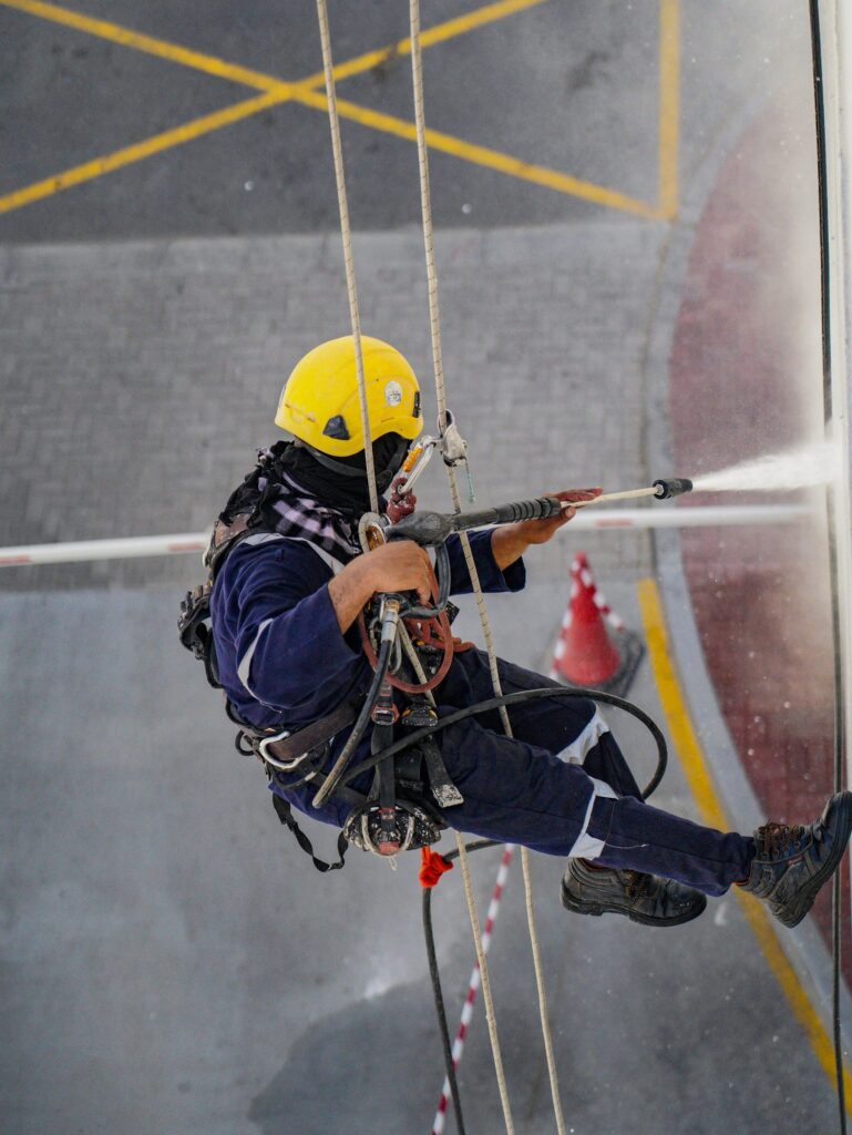A rope access technician in helmet and harness washing a hull with a pressure lance