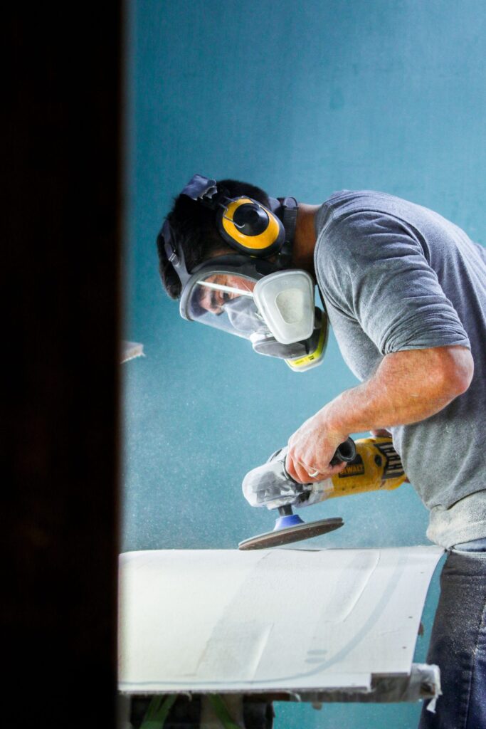 A worker in a mask and ear defenders sanding a surface before it is coated
